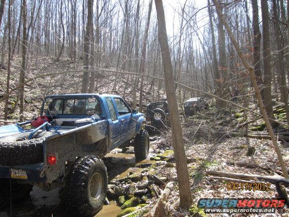 Blue pickup truck and other off-road vehicles navigating a muddy, rocky creek in a forest
