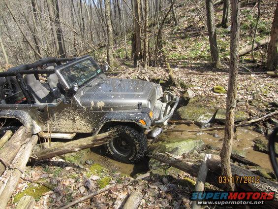 Gray Jeep Wrangler navigating a muddy creek bed with logs