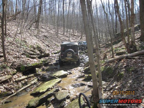 Two Jeeps navigating a muddy, rocky stream in a wooded area. Date stamp reads 03/17/2012