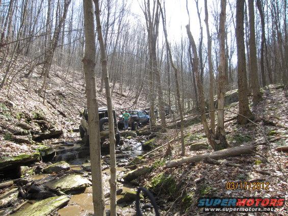 two off-road vehicles driving through a rocky creek in a forest