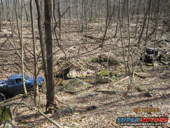 Blue pickup truck and a dark SUV off-roading in a forest with a stream