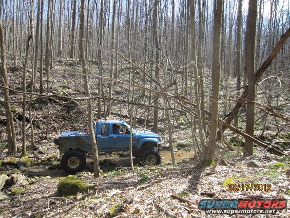 Blue pickup truck with large tires driving through a muddy stream in a dense forest