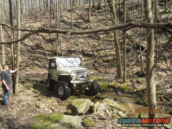 White Jeep Wrangler with large tires driving through a rocky stream bed in a forest. A person stands to the left