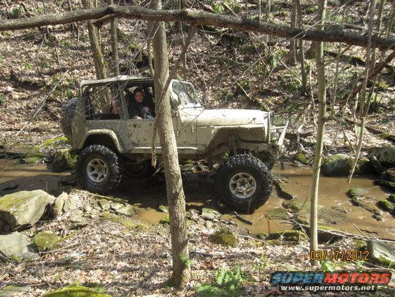 Mud-covered Jeep Wrangler driving through a shallow creek in a wooded area