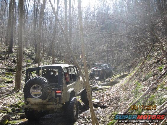 Two off-road vehicles navigate a rocky, wooded trail