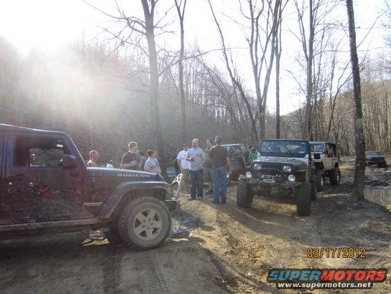 Several Jeeps parked on a muddy dirt road in a wooded area. People are standing around the vehicles