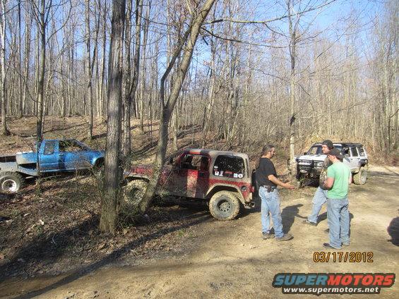 Red Jeep Wrangler and blue pickup truck parked on a dirt path in a forest with people standing around