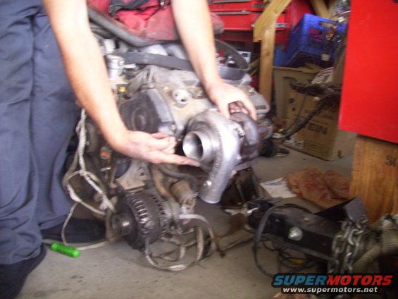Engine block with turbocharger being held by hands. Green screwdriver on floor