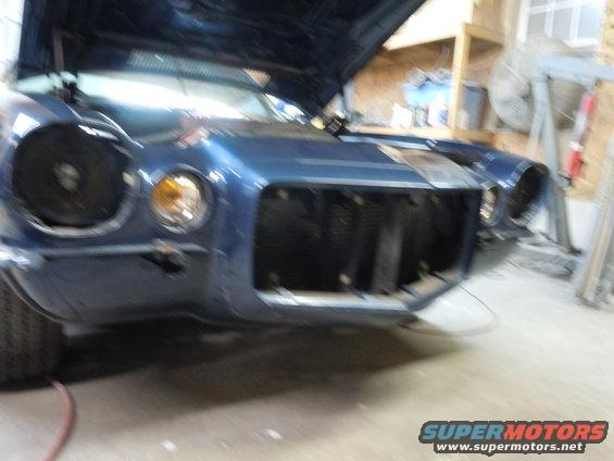 Blue coupe, hood up, front end disassembled. Grille missing, headlight assembly visible. Supermotors.net logo at bottom right