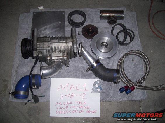 Supercharger assembly with hoses, belts, and mounting plate. Paper note reads 'MAC1 3-18-12 PROBE TALK CLUB PROTEGE PERFORMANCE PROBE'