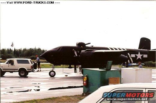 White SUV parked next to a large airplane covered in black tarp. A person is standing by the SUV