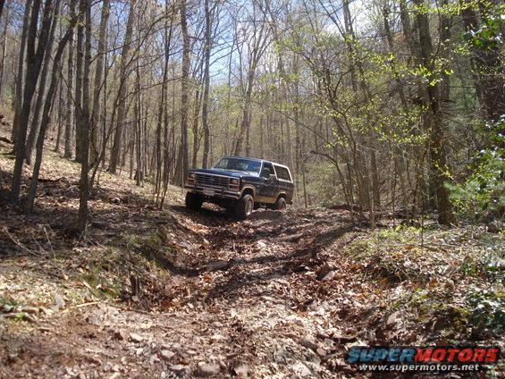 1985 Ford Bronco driving on a muddy, wooded trail