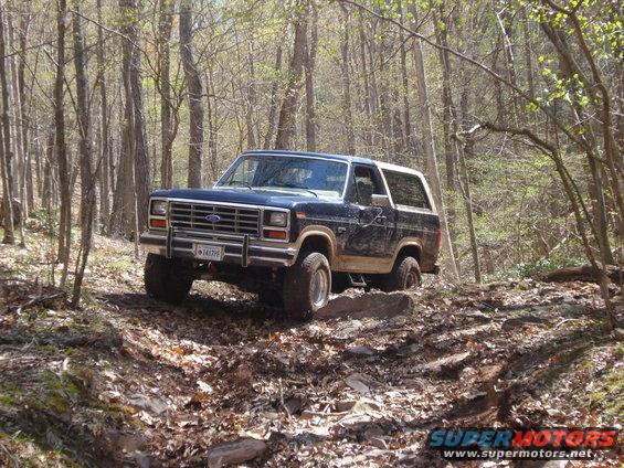 Blue Ford Bronco with white top driving on a dirt trail in a wooded area. License plate is visible