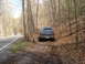 1985 Ford Bronco - A blue Ford Bronco is parked on a dirt path beside a winding road through a wooded area.