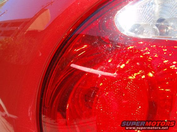 Red car tail light with visible scratches and debris. Watermark reads SUPERMOTORS.NET