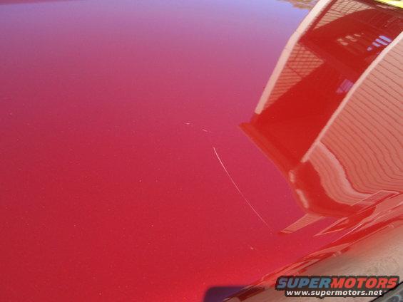 Red car hood with a prominent scratch and reflection of a building