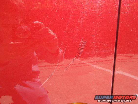 Red car door with multiple white scratches and a reflection of a person holding a camera