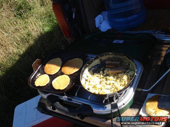 Camp stove cooking pancakes and sausage with a cooler and water jug nearby