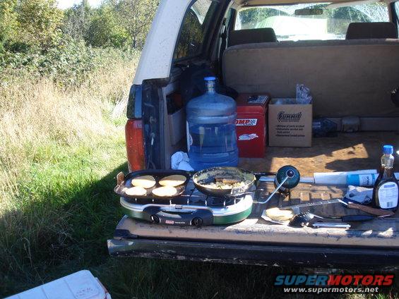 Tailgate of a vehicle with a portable stove cooking pancakes, a large water jug, and other supplies