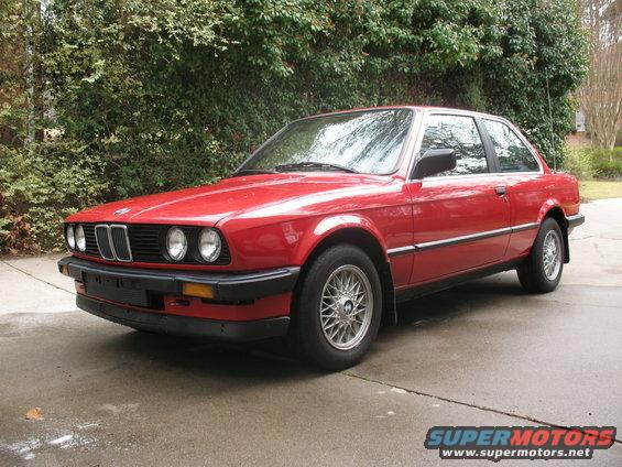 Red 1986 BMW 3 Series coupe parked on a driveway, showing its front-left quarter. The car has a black grille, round headlights, and silver multi-spoke wheels