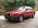 1986 BMW 3 Series - A red 1986 BMW 3 Series coupe is viewed from the front-left, parked outdoors on a paved surface with trees in the background.