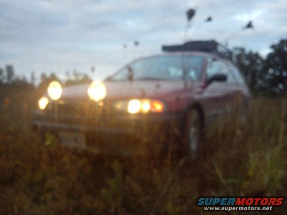 red wagon with roof rack and multiple spotlights, parked in a grassy field at dusk