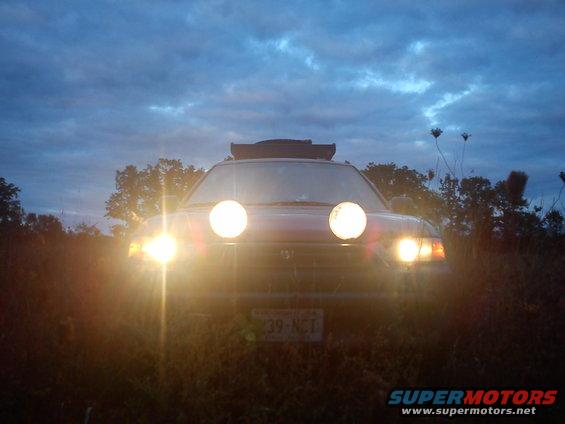 Subaru Outback with two large round auxiliary lights mounted on the front, facing the camera. It has a roof rack and is parked outdoors at dusk
