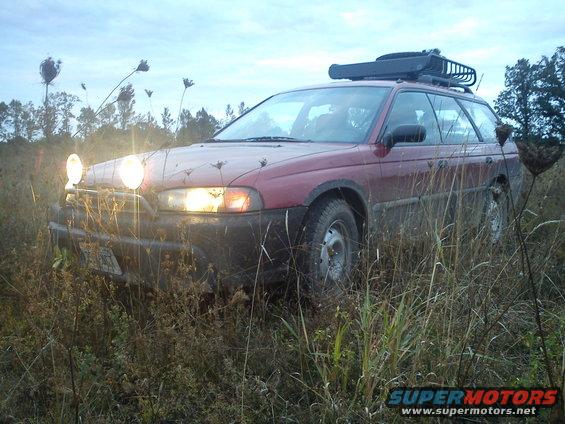 Red 1998 Subaru Legacy wagon with roof rack and auxiliary lights parked in tall grass