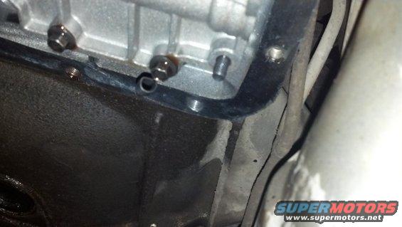 Transmission pan gasket with bolts and oil residue. Part of a vehicle's undercarriage is visible