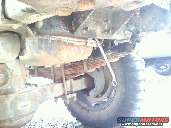 Undercarriage of a large truck showing leaf springs, axle, and a wrench. Another vehicle is partially visible in the background