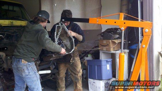 two guys using an engine hoist to lift a transmission out of a yellow vehicle