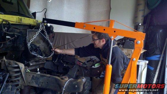Yellow vehicle chassis with engine removed, being worked on by a man using an orange engine hoist