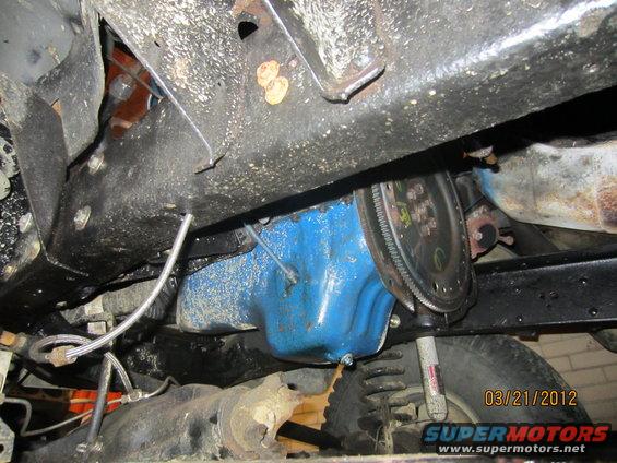 Underside view of a vehicle's transmission bell housing and oil pan. Shows part of the frame and suspension