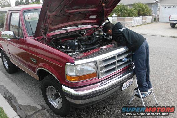 Man on step stool looking at engine of red Ford Bronco with hood up. License plate visible