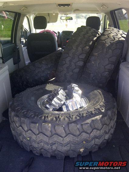 Several large off-road tires and chrome lug nuts piled in the back of a vehicle