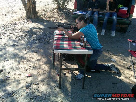 Truck bed with people sitting, a woman kneels at a table with a rifle, and a folding chair