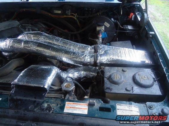 Engine bay with insulated intake pipes and battery. Some warning labels are visible