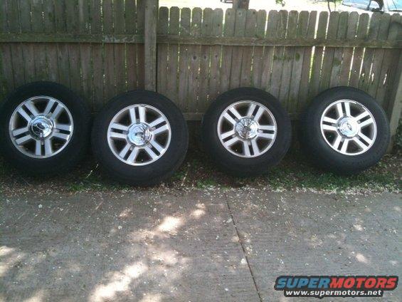 Four alloy wheels with tires lined up against a wooden fence