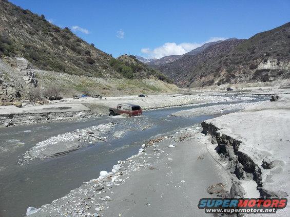 Red SUV driving through a shallow river in a canyon with other vehicles in the background