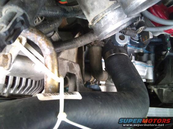 Close-up of engine bay with hoses, clamps, and some metal components visible. A barcode tag is attached with zip ties
