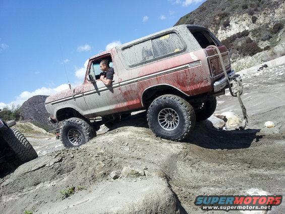 1978 Ford Bronco climbing a dirt hill with a driver inside. Another vehicle's tire is visible on the left