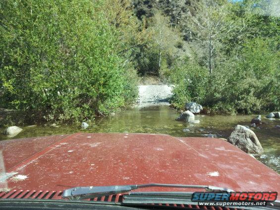 Red vehicle hood and windshield wipers in foreground, crossing a shallow stream with trees and rocks