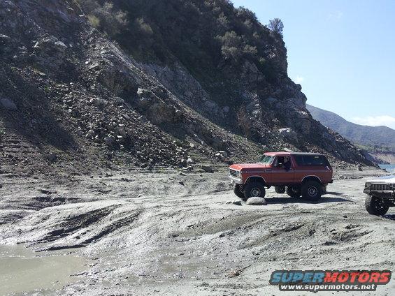 Red 1978 Ford Bronco driving on a muddy, rocky shore