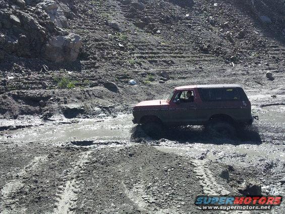 Red 1978 Ford Bronco driving through a muddy track, side view