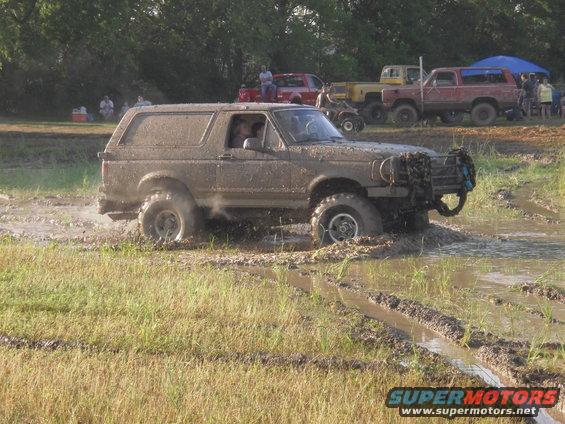 Muddy SUV driving through a mud pit with other vehicles and people in the background
