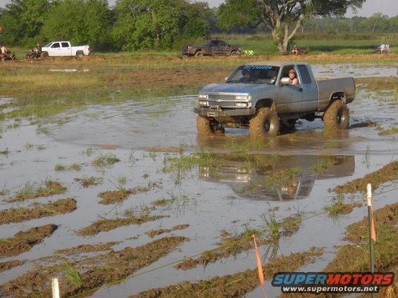 Blue pickup truck with lifted suspension driving through mud, other vehicles in background