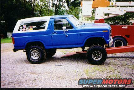 1985 Ford Bronco, blue with white top, on a red trailer. Several stickers on the rear passenger window
