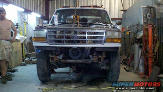 White Ford truck with front end disassembled, lifted by a chain hoist