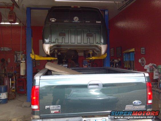 Rear view of a dark green Ford F-250 Super Duty truck, with another truck lifted above it in a garage