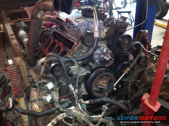 Engine block with pistons exposed, surrounded by hoses and wiring in a garage